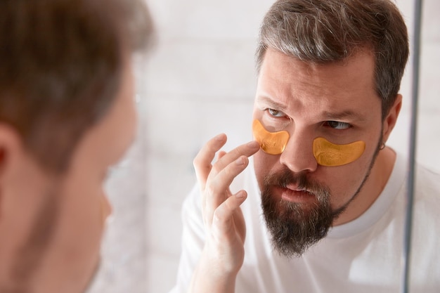 Ritratto di uomo in maglietta bianca che applica bende per gli occhi sul viso in bagno. Procedura mattutina per la cura di sé. Concetto di trattamento di bellezza