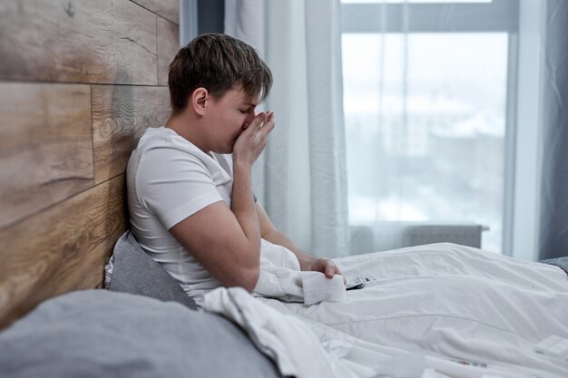 Ritratto di uomo malsano che sente dolore alla gola e febbre alta, seduto sul letto da solo. Coronavirus e COVID-19. concetto di mal di testa, malattia, influenza, medicina e sanità