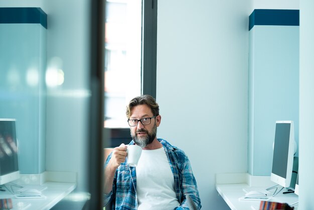 Ritratto di uomo maturo che guarda la telecamera mentre beve caffè in ufficio - lavoro online freelance con stile di vita concetto di computer - persone caucasiche adulte nello spazio di coworking - umore blu colori persone moderne