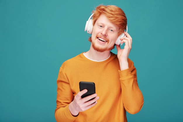 Ritratto di uomo positivo bello giovane rossa con la barba che regola le cuffie senza fili mentre si sceglie la traccia musicale sullo smartphone