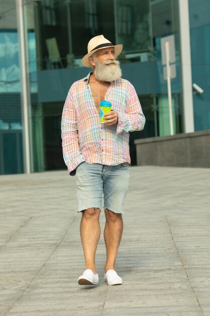 Ritratto di uomo positivo senior barbuto hipster, avendo pausa caffè all'aperto su contesti urbani.