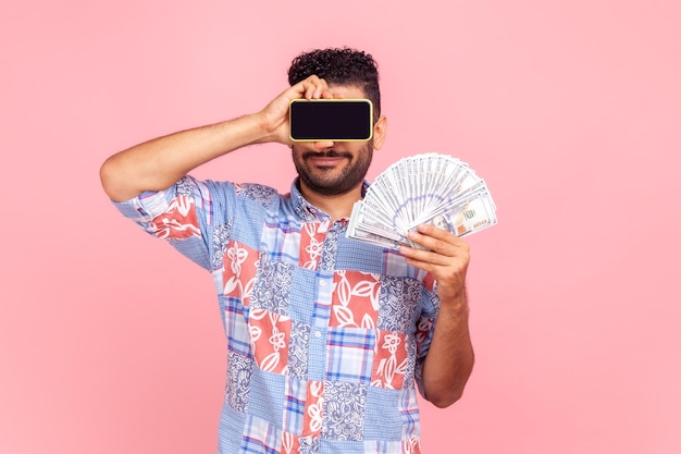 Ritratto di uomo sconosciuto con la barba che indossa una camicia blu in stile casual in posa con banconote in dollari e che copre gli occhi con uno smartphone con schermo vuoto. Studio indoor girato isolato su sfondo rosa.