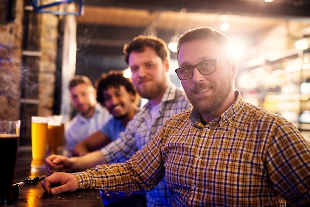 Ritratto di uomo sorridente bello seduto al bar con gli amici. Bicchieri di birra alla spina e fumo della sigaretta di fronte.