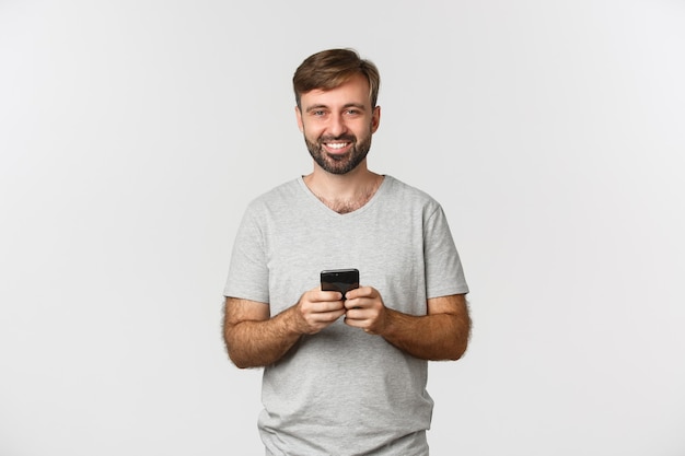 Ritratto di uomo sorridente felice con la barba, indossa la maglietta, utilizzando il telefono cellulare, in piedi su bianco