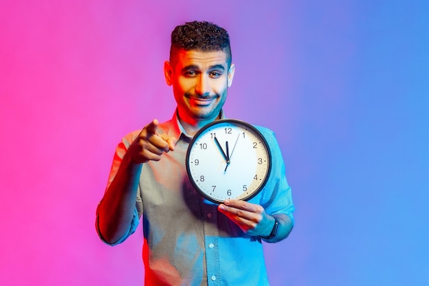 Ritratto di uomo sorridente in camicia che punta il dito verso la telecamera tenendo in mano un grande orologio da parete, tempo di azione, motivazione. Studio indoor girato isolato su sfondo colorato con luce al neon.