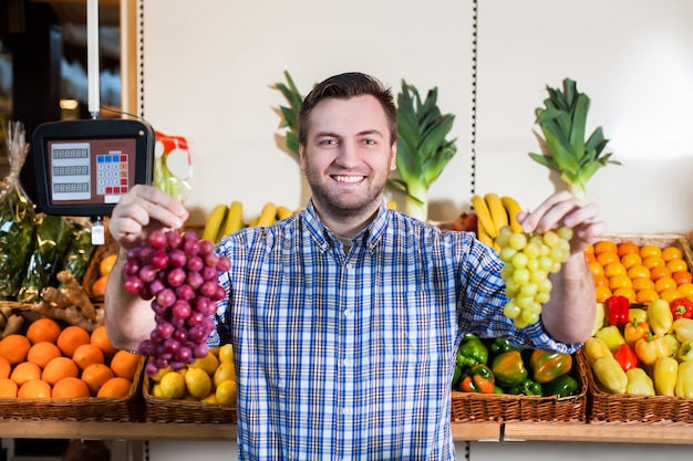 Ritratto di uomo sorridente in camicia che vende uva matura in negozio. Scatole con frutta e verdura