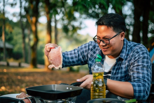 Ritratto di uomo viaggiatore asiatico occhiali bistecca di maiale frittura barbecue in padella o pentola per arrostire in un campeggio concetto di stile di vita campeggio viaggio cucina all'aperto