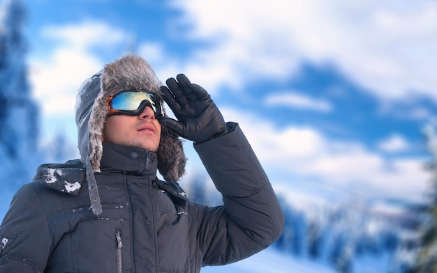 Ritratto di vacanza invernale di un uomo che indossa un cappello paraorecchie e occhiali da sci con le montagne