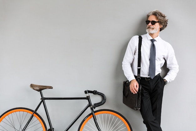 Ritratto di vecchio uomo d'affari alla moda in occhiali da sole che guarda da parte mentre stando con la bicicletta sopra la parete grigia all'aperto