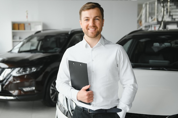 Ritratto di venditore professionista di mentalità aperta in showroom di automobili uomo caucasico in camicia bianca formale si trova accanto a un'auto lussuosa e guarda la fotocamera