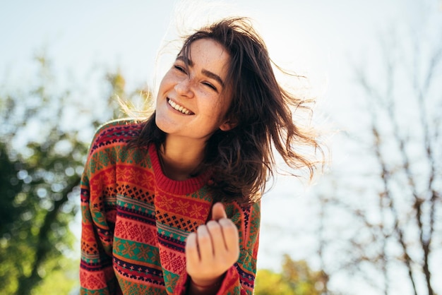 Ritratto di vista dal basso di felice bella giovane donna sorridente ampiamente con i capelli ventosi e le lentiggini godendo il caldo clima autunnale che indossa un maglione in posa sullo sfondo della luce solare della natura