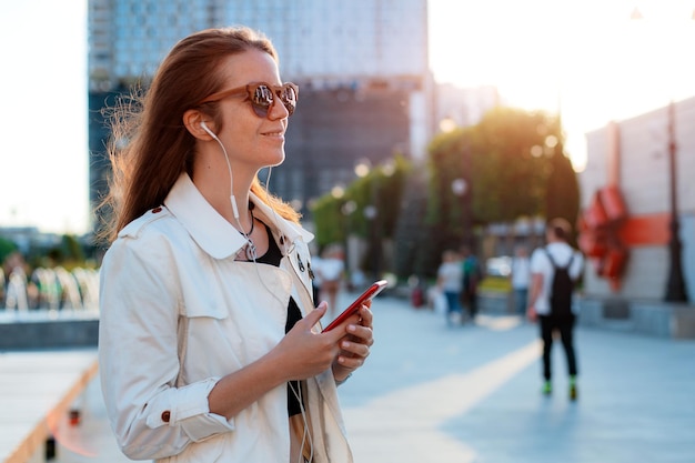 Ritratto di vista frontale moda moderna donna felice hipster a piedi e utilizzando uno smart phone su una strada cittadina indossando occhiali da sole al sole estivo