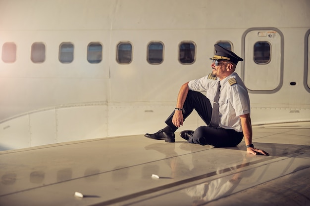 Ritratto di vista laterale a tutta lunghezza di un bell'uomo in uniforme da lavoro seduto sull'ala di un aereo passeggeri sullo sfondo della fusoliera