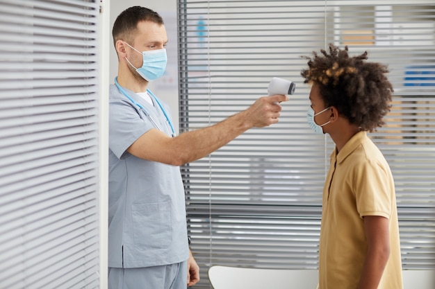 Ritratto di vista laterale del medico maschio che controlla la temperatura del ragazzo afro-americano che indossa la maschera mentre aspetta in fila presso la clinica medica, copia spazio