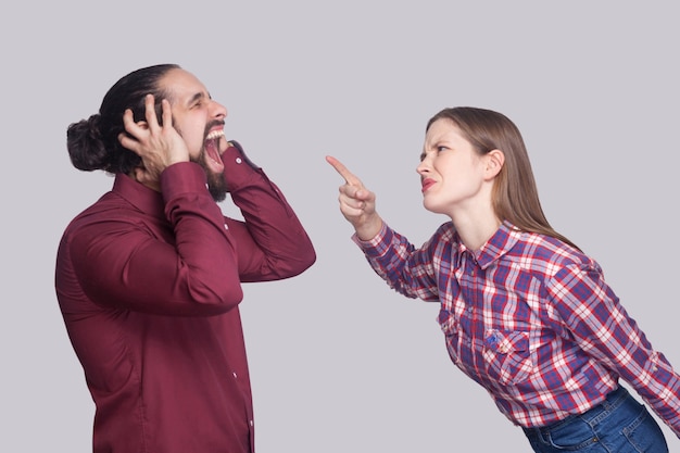 Ritratto di vista laterale del profilo di una donna arrabbiata in piedi e che incolpa di urlare un uomo infelice con i capelli neri raccolti. girato in studio al coperto, isolato su sfondo grigio.