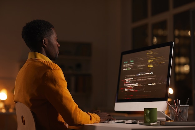 Ritratto di vista laterale dell'uomo afroamericano contemporaneo che esamina lo schermo del computer mentre lavora a tarda notte il codice di scrittura, lo spazio della copia
