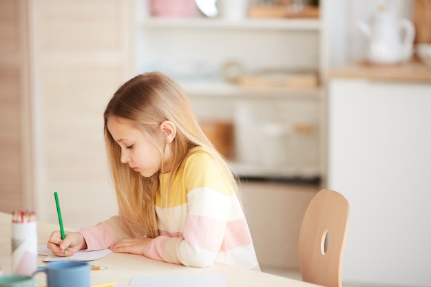 Ritratto di vista laterale della bambina sveglia disegnare immagini o fare i compiti mentre è seduto al tavolo in interni domestici, copia dello spazio