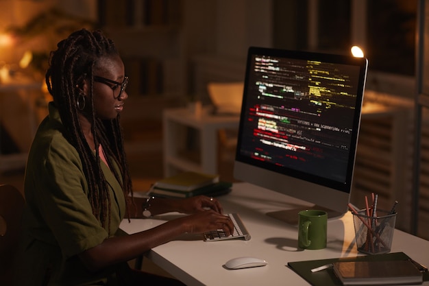 Ritratto di vista laterale della donna afroamericana contemporanea che scrive codice e che guarda lo schermo del computer mentre lavora nell'ufficio scuro, spazio della copia