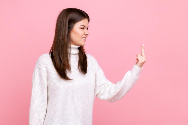 Ritratto di vista laterale della donna aggressiva in piedi e mostra il segno del cazzo, esprimendo emozioni negative, indossando un maglione bianco in stile casual. Studio indoor girato isolato su sfondo rosa.