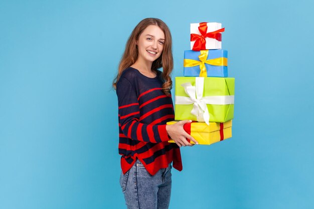 Ritratto di vista laterale della donna che indossa un maglione casual a righe in piedi con una pila di regali nelle mani che tengono scatole regalo guardando sorridente alla telecamera Studio indoor girato isolato su sfondo blu