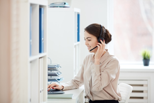 Ritratto di vista laterale della donna di affari sorridente che parla al microfono mentre lavora con il computer portatile nell'interiore dell'ufficio