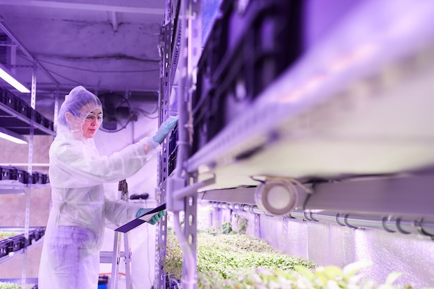 Ritratto di vista laterale dello scienziato femminile che tiene appunti mentre lavora nella serra della scuola materna o nel laboratorio della piantagione, spazio della copia