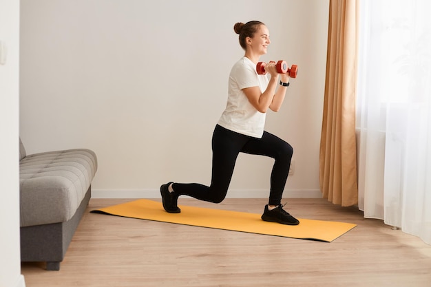 Ritratto di vista laterale di donna che fa affondo per il suo obiettivo bella ragazza in maglietta bianca e leggins neri che sollevano manubri al chiuso su un tappetino da yoga Stile di vita sano