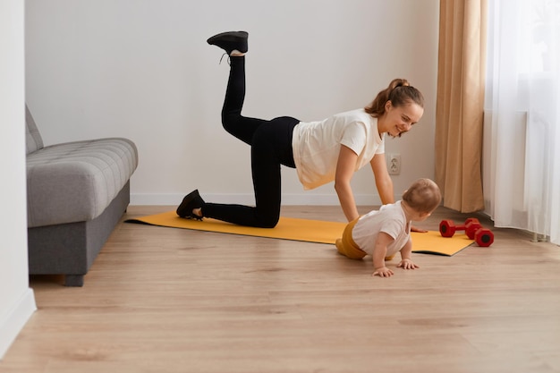 Ritratto di vista laterale di donna in forma che fa esercizio di calcio d'asino piegato al ginocchio in posizione a quattro zampe allenando i glutei a casa con la sua piccola figlia neonata, fitness e stile di vita sano.