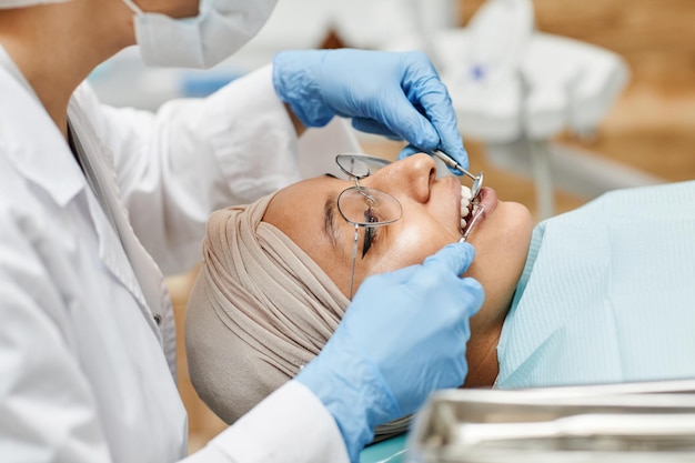 Ritratto di vista laterale di giovane donna araba sdraiata sulla poltrona del dentista con strumenti di detenzione del dentista durante l'esame...