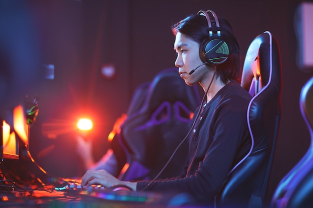 Ritratto di vista laterale di giovane pro-giocatore asiatico che gioca ai videogiochi in camera oscura, copia dello spazio