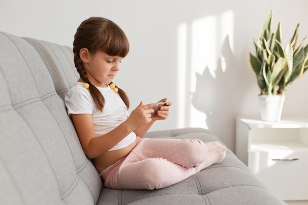 Ritratto di vista laterale di piccola bambina dai capelli scuri concentrata che indossa una maglietta casual bianca utilizzando il telefono cellulare seduto sul divano, giocando ai videogiochi.