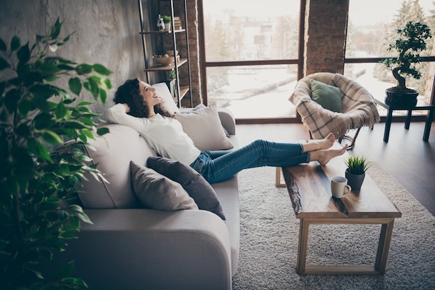 Ritratto di vista laterale di Profille della ragazza allegra allegra affascinante che si siede sul sofà che riposa trascorre la giornata al soggiorno in stile interno moderno loft industriale al chiuso