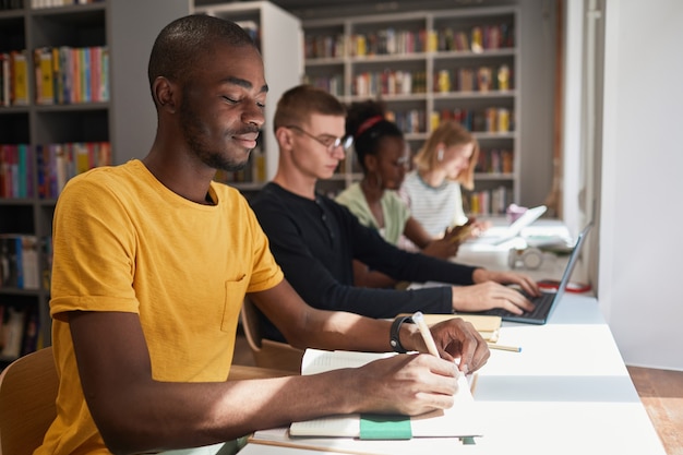 Ritratto di vista laterale di un gruppo eterogeneo di studenti seduti in fila mentre studiano nella copia della biblioteca scolastica...