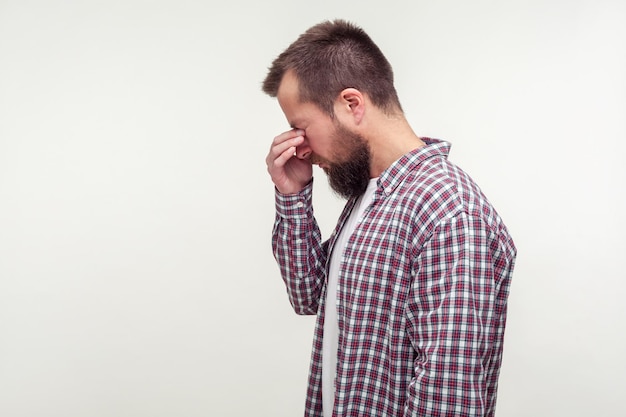 Ritratto di vista laterale di un uomo barbuto infelice in camicia a quadri casual in piedi con la testa in giù, stropicciandosi gli occhi e piangendo, stanco umore depresso. posto vuoto per testo, copia spazio. isolato su sfondo bianco