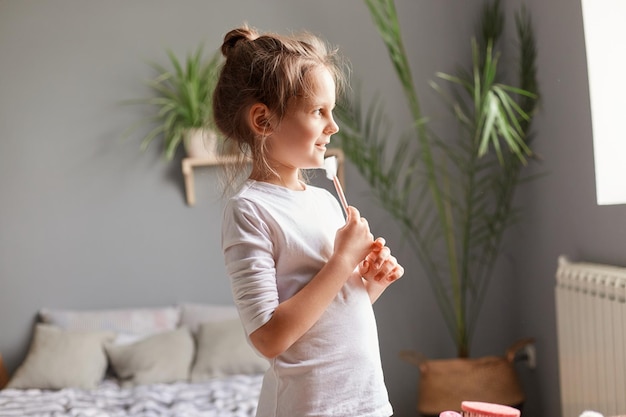 Ritratto di vista laterale di una bambina dai capelli castani carina che indossa una camicia bianca che tiene un pennello cosmetico per il trucco che guarda avanti in piedi nell'interno della casa della camera da letto