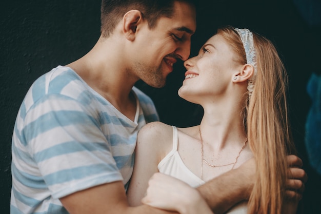 Ritratto di vista laterale di una bella giovane coppia che abbraccia e sorride prima di baciarsi contro un muro nero all'aperto.
