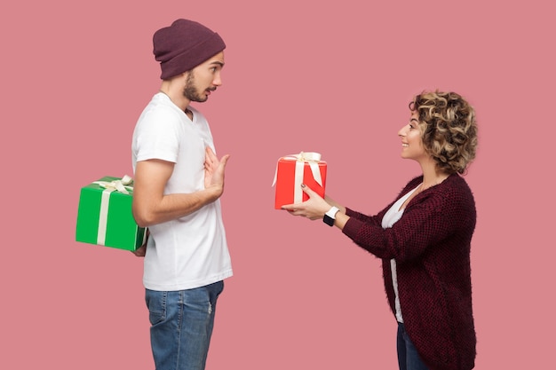 Ritratto di vista laterale di una coppia incredibile di amici in piedi in stile casual, ragazza che dà una scatola regalo, ragazzo che si nasconde dietro un regalo, festeggia l'anniversario. Isolato, interno, girato in studio, sfondo rosa