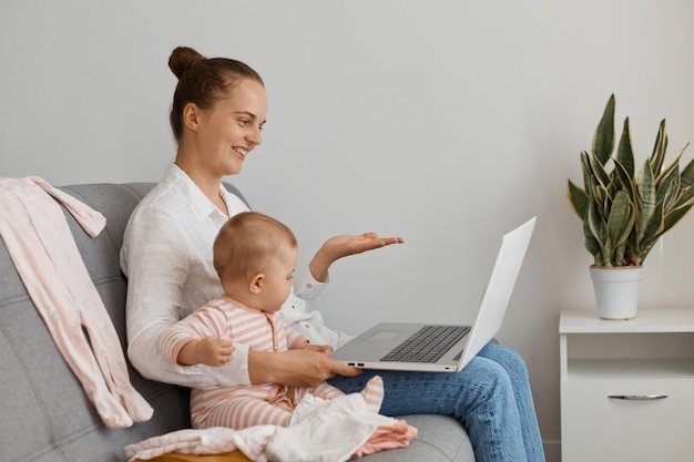 Ritratto di vista laterale di una donna che indossa una camicia bianca e jeans seduta sul divano con la figlia piccola e fa una videochiamata sul laptop, allargando la mano, blogger che parla con i follower.