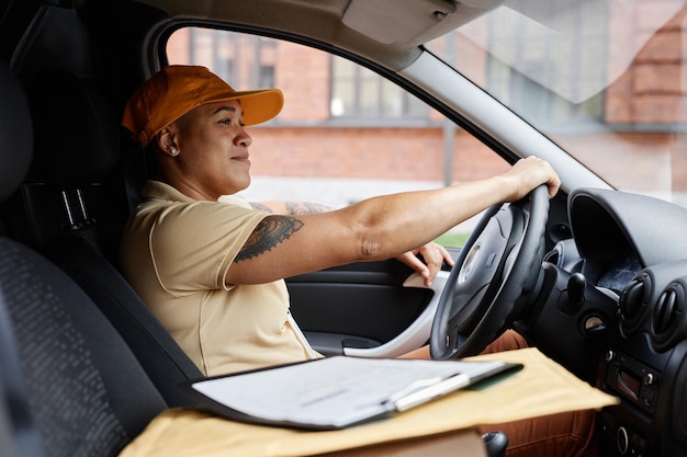 Ritratto di vista laterale di una giovane donna multietnica che guida un camion per le consegne e indossa l'uniforme