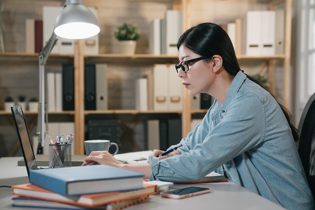 Ritratto di vista laterale interna di una donna giapponese asiatica impegnata intelligente che prende appunti mentre è seduto a casa con un computer portatile. vista laterale ragazza del college intelligente alla ricerca di informazioni notebook online facendo i compiti