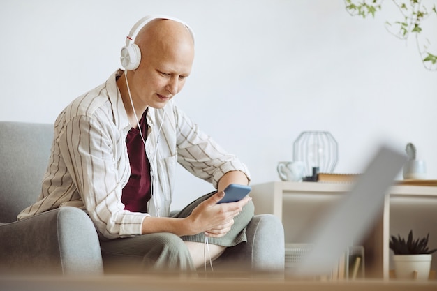 Ritratto di vista laterale minima di donna adulta calva che indossa le cuffie durante l'ascolto di musica tramite smartphone in interni domestici accoglienti, alopecia e consapevolezza del cancro, spazio di copia