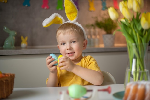 Ritratto emotivo di un ragazzino allegro che indossa orecchie da coniglio il giorno di Pasqua che ride allegramente gioca con colorate uova di Pasqua seduto a un tavolo in cucina