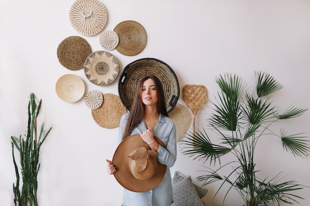 Ritratto estivo di una giovane donna graziosa in un vestito e con in mano un cappello in interni esotici boho