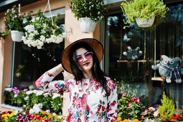 Ritratto estivo di una ragazza bruna con occhiali rosa e cappello contro il negozio di fiori