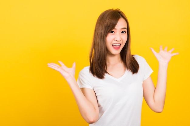 Ritratto felice asiatico bella giovane donna carina adolescente in piedi indossare t-shirt Sorpreso eccitato urlando bocca aperta mostra la mano che guarda alla macchina fotografica isolata, girato in studio su sfondo giallo con spazio di copia