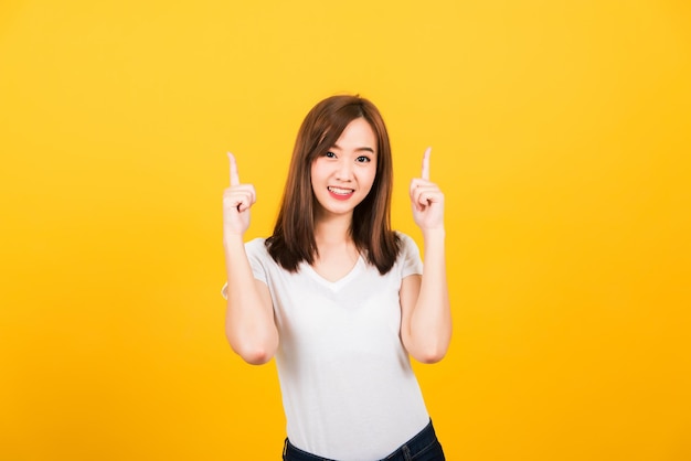 Ritratto felice asiatico bella giovane donna carina adolescente indossare t-shirt in piedi fa gesto due dita puntano verso l'alto sopra guardando alla fotocamera isolata, girato in studio su sfondo giallo con spazio di copia