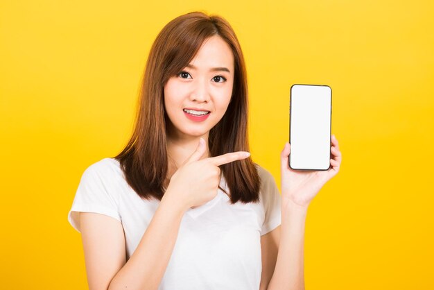 Ritratto felice asiatico bella giovane donna carina sorriso in piedi indossare t-shirt facendo il dito puntato sullo schermo vuoto dello smartphone guardando alla fotocamera isolata, studio girato sfondo giallo con spazio di copia