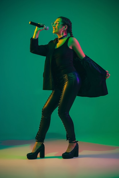 Ritratto femminile caucasico della cantante isolato sulla parete verde alla luce al neon. Bellissimo modello femminile in abbigliamento nero con microfono. Concetto di emozioni umane, espressione facciale, pubblicità, musica, arte.