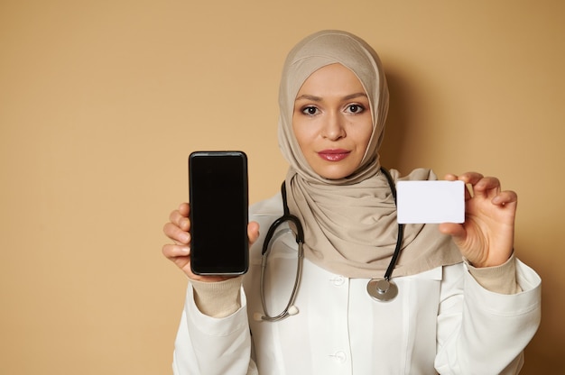 Ritratto fiducioso di una giovane dottoressa musulmana che indossa l'hijab che tiene una carta in bianco di plastica bianca e uno smartphone e li mostra alla parte anteriore