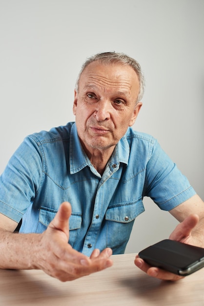 Ritratto fotografico autentico di uomo anziano che parla seduto a tavola con il telefono cellulare in mano, moderno anziano candido avanzato su sfondo bianco